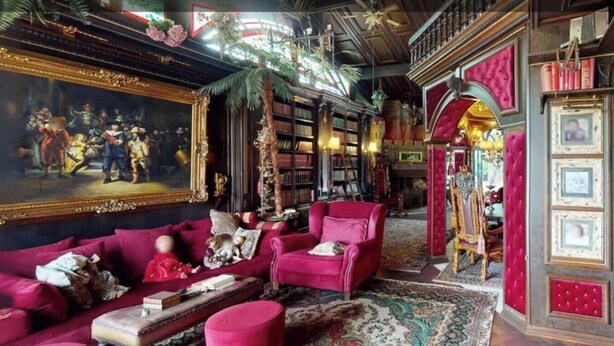 A room with a large classical painting, a red sofa, a bookshelf, and a red armchair.