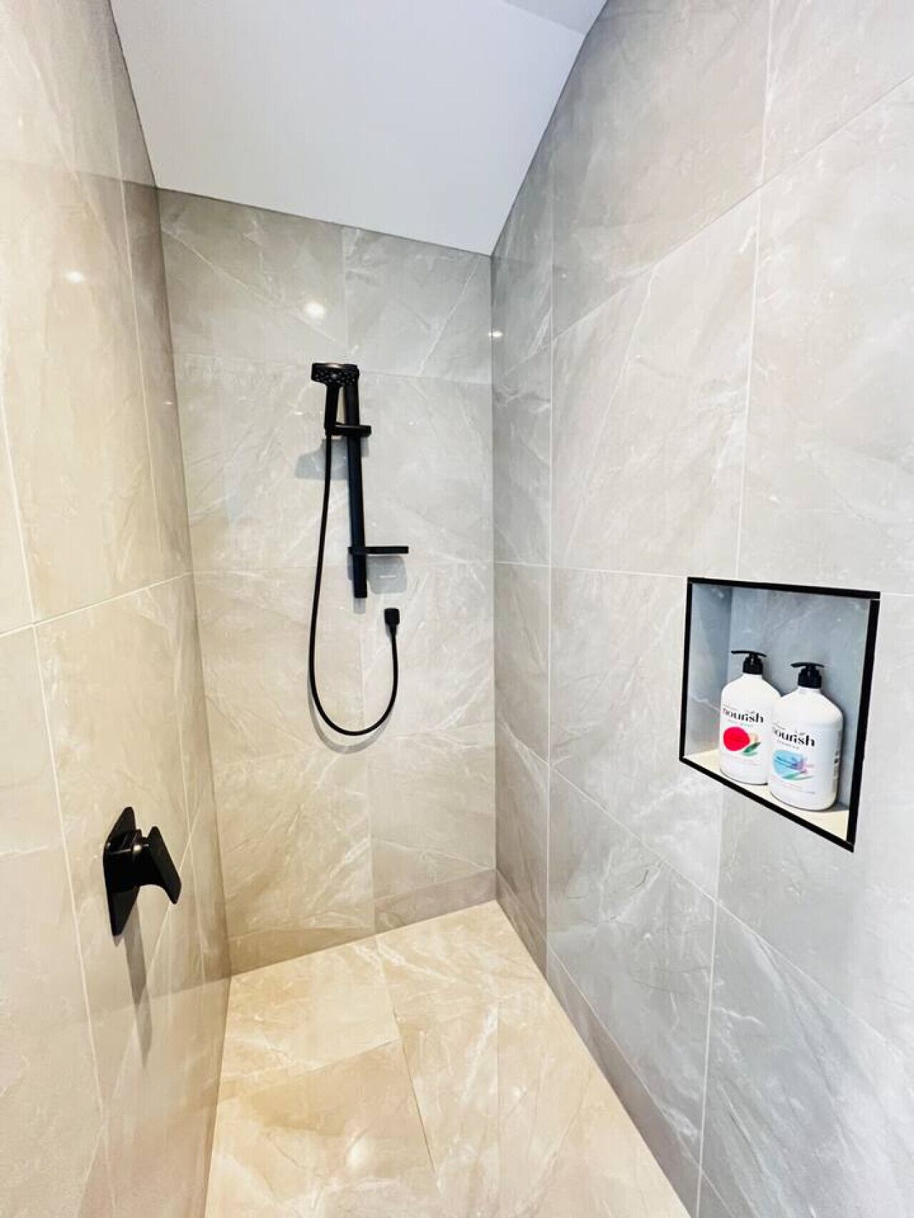 Une douche moderne dotée d’un pommeau de douche à main, d’un panneau de commande mural et d’une étagère contenant deux flacons de gel douche.