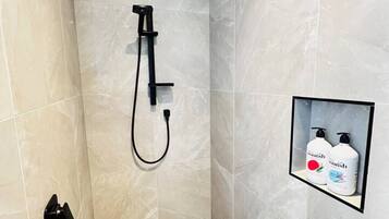 Une douche moderne dotée d’un pommeau de douche à main, d’un panneau de commande mural et d’une étagère contenant deux flacons de gel douche.