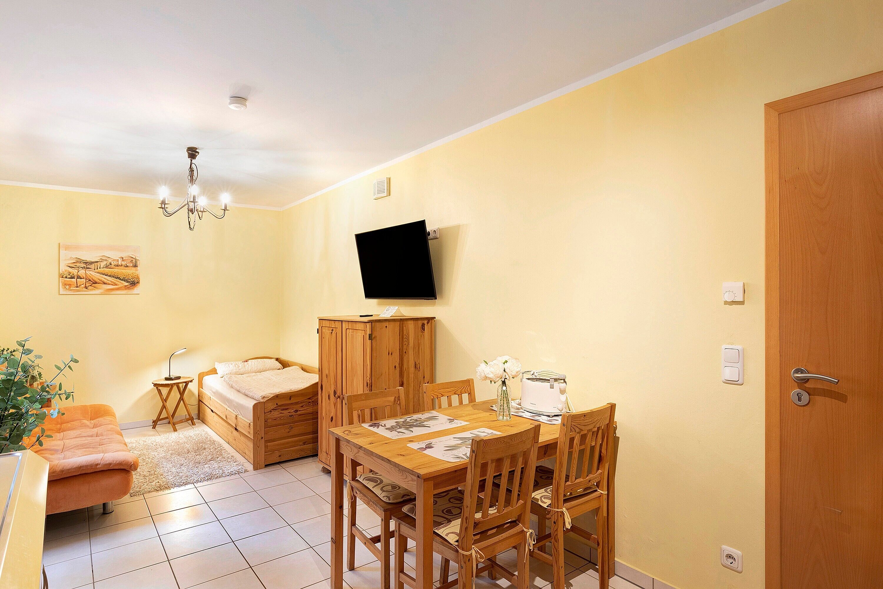 A room with a bed, a dining table with chairs, a TV mounted on the wall, and a door.