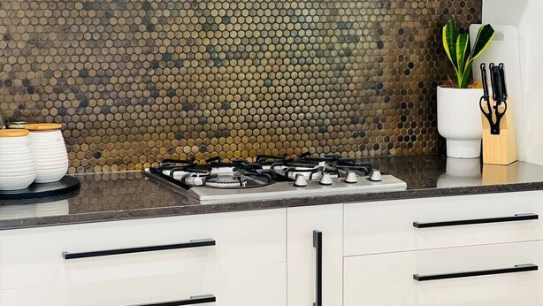Una cucina con piano cottura a gas, mobili bianchi, zoccolo a mosaico e una pianta in vaso.