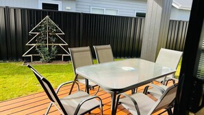 Una veranda esterna con un tavolo in vetro e sedie in metallo, un pontile in legno, una recinzione nera e una struttura a forma di triangolo per piante.