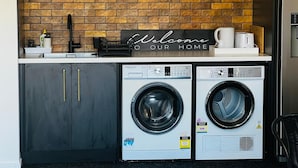 A laundry area with two front-loading washing machines, a coffee maker, and a 'Welcome to Our Home' sign on a brick wall.