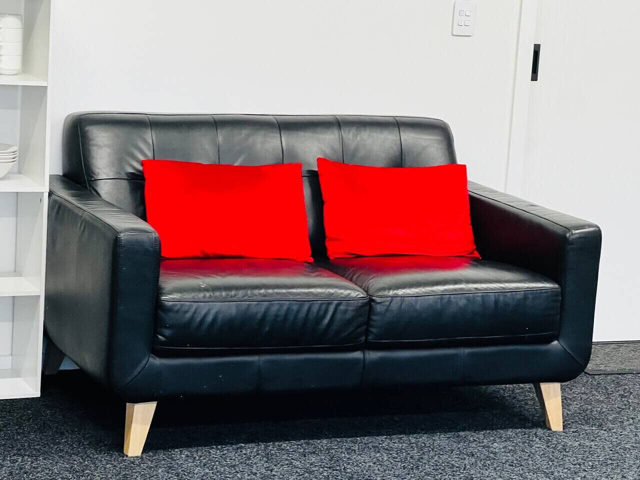 A black leather sofa with two red cushions, wooden legs, and a carpeted floor.