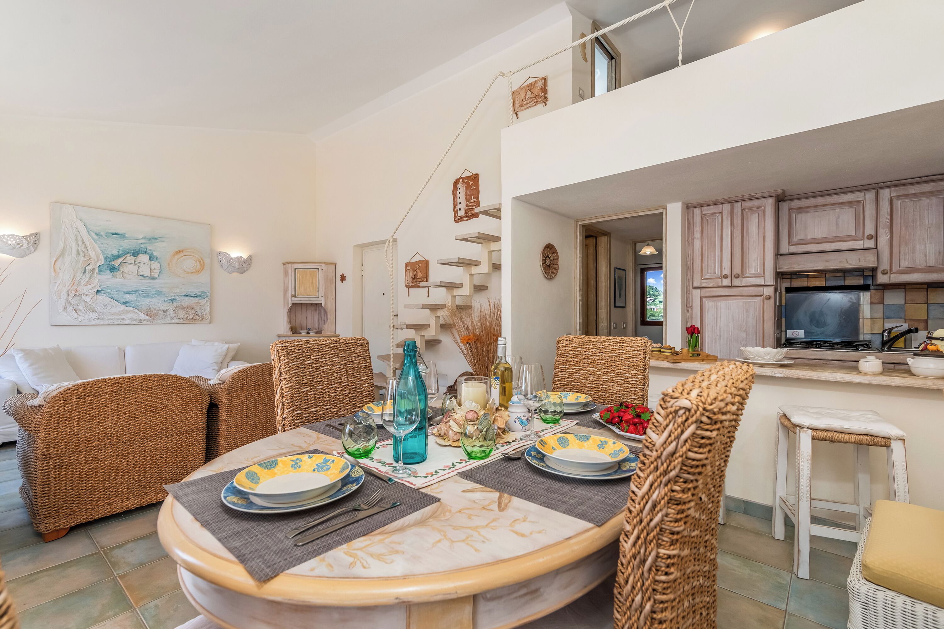 Uma sala de jantar com mobiliário de vime, uma mesa posta para quatro pessoas e uma cozinha ao fundo.