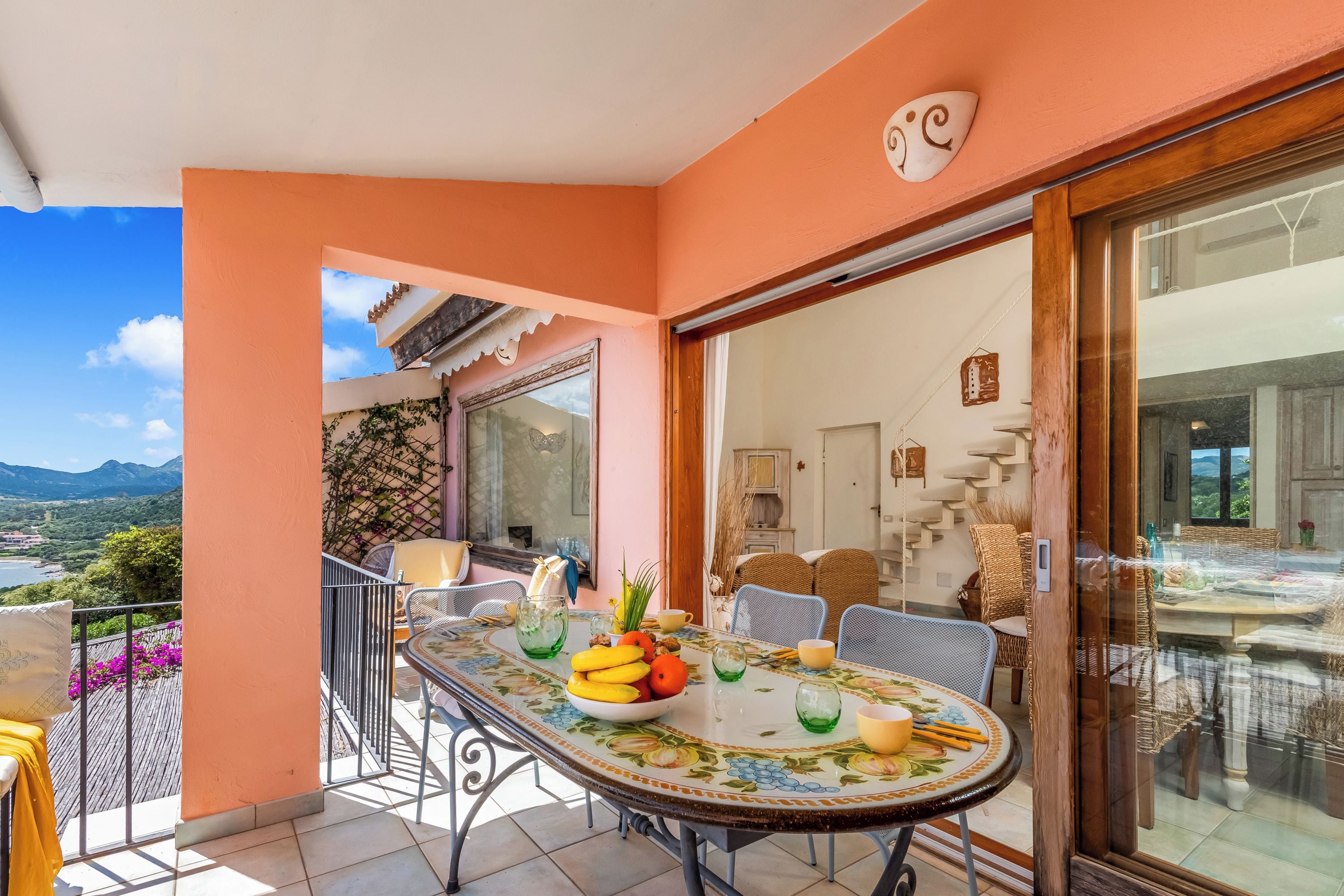 Un balcon avec une table à manger dressée, ornée de fruits, des chaises et une vue sur le paysage.