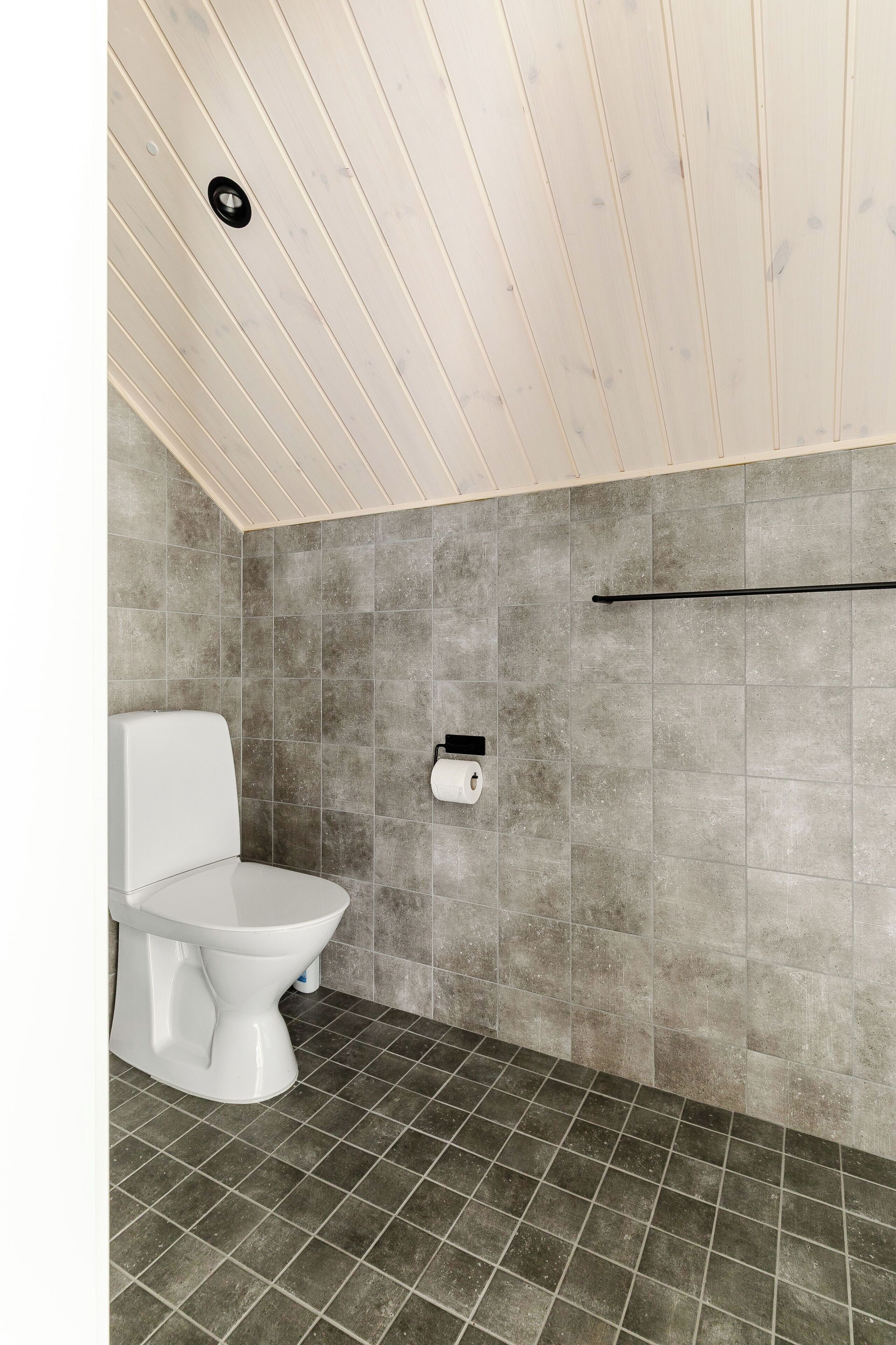 A bathroom with a white toilet, grey tiled walls, and a sloped ceiling.