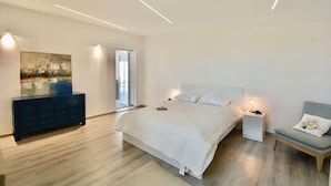 A modern bedroom with a large bed, bedside tables, a chair, a painting, and a sliding glass door leading to a balcony. - Rari House by Geco Vacation Rentals (Brucoli)