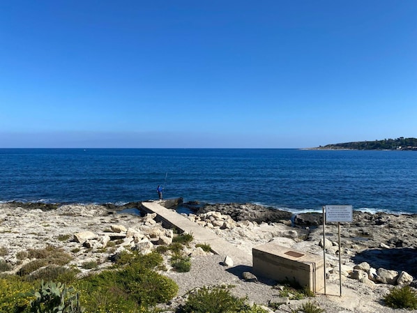 A rocky shoreline with a clear view of the sea and a sign indicating a protected area. - Rari House by Geco Vacation Rentals (Brucoli)