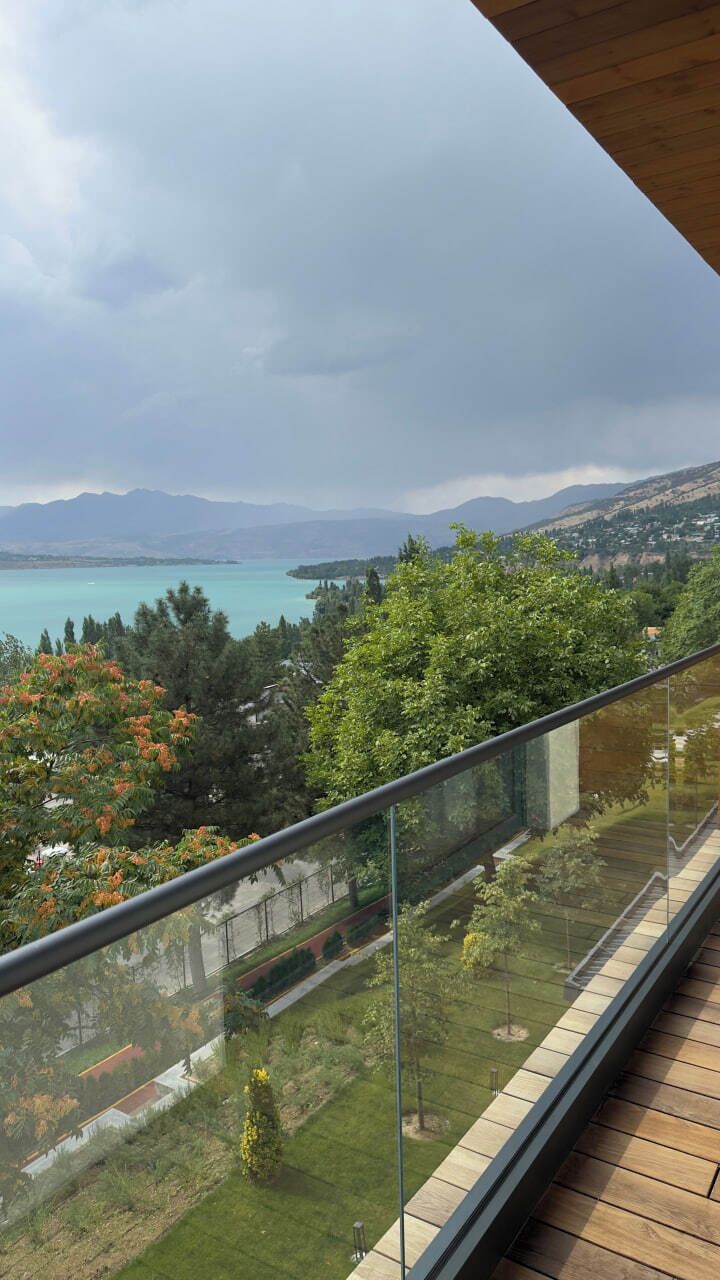 A balcony with a glass railing overlooking a landscaped garden and a distant view of a lake and mountains.