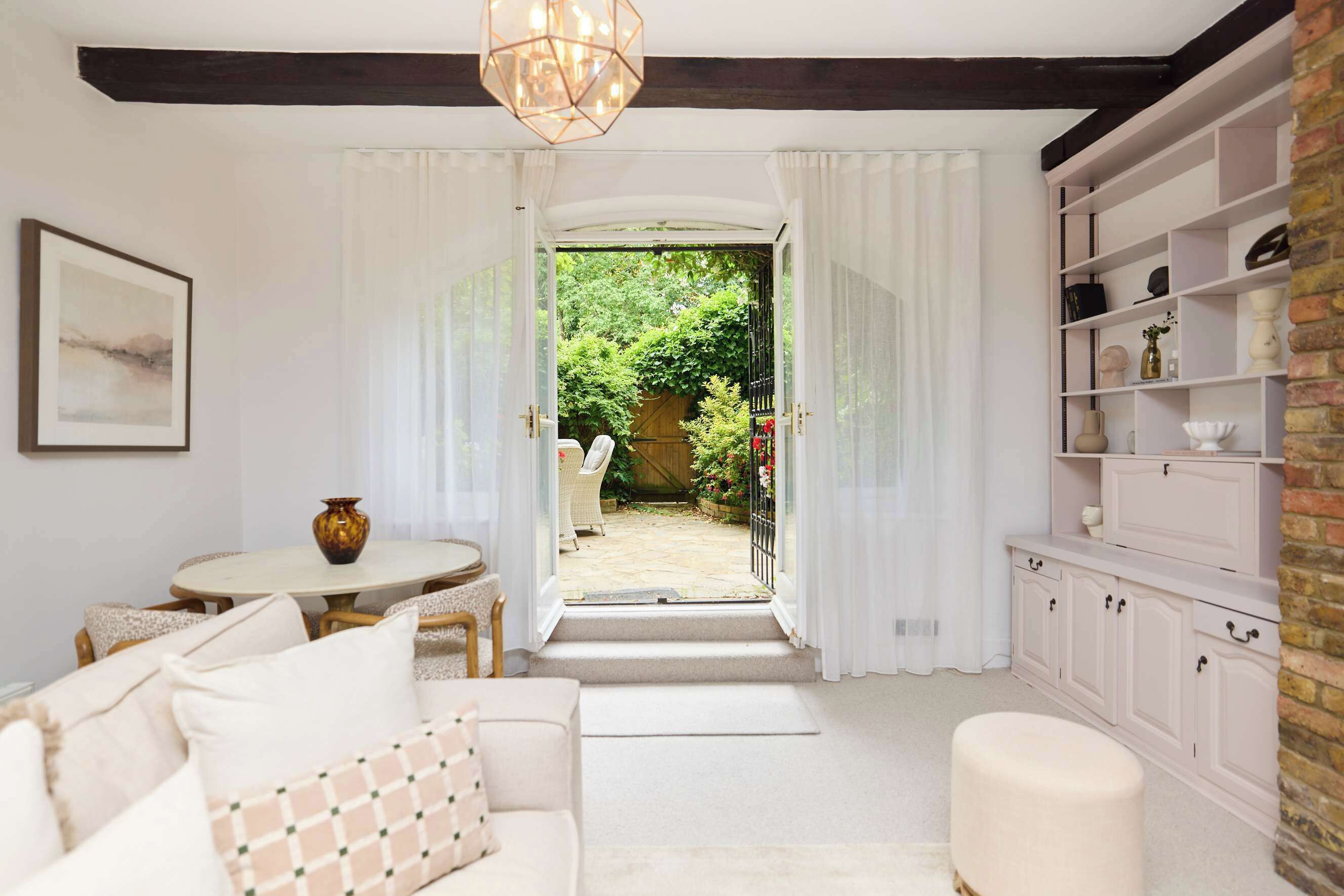A room with a white sofa, a dining table with chairs, a white cabinet, and a view of a garden through an open door.