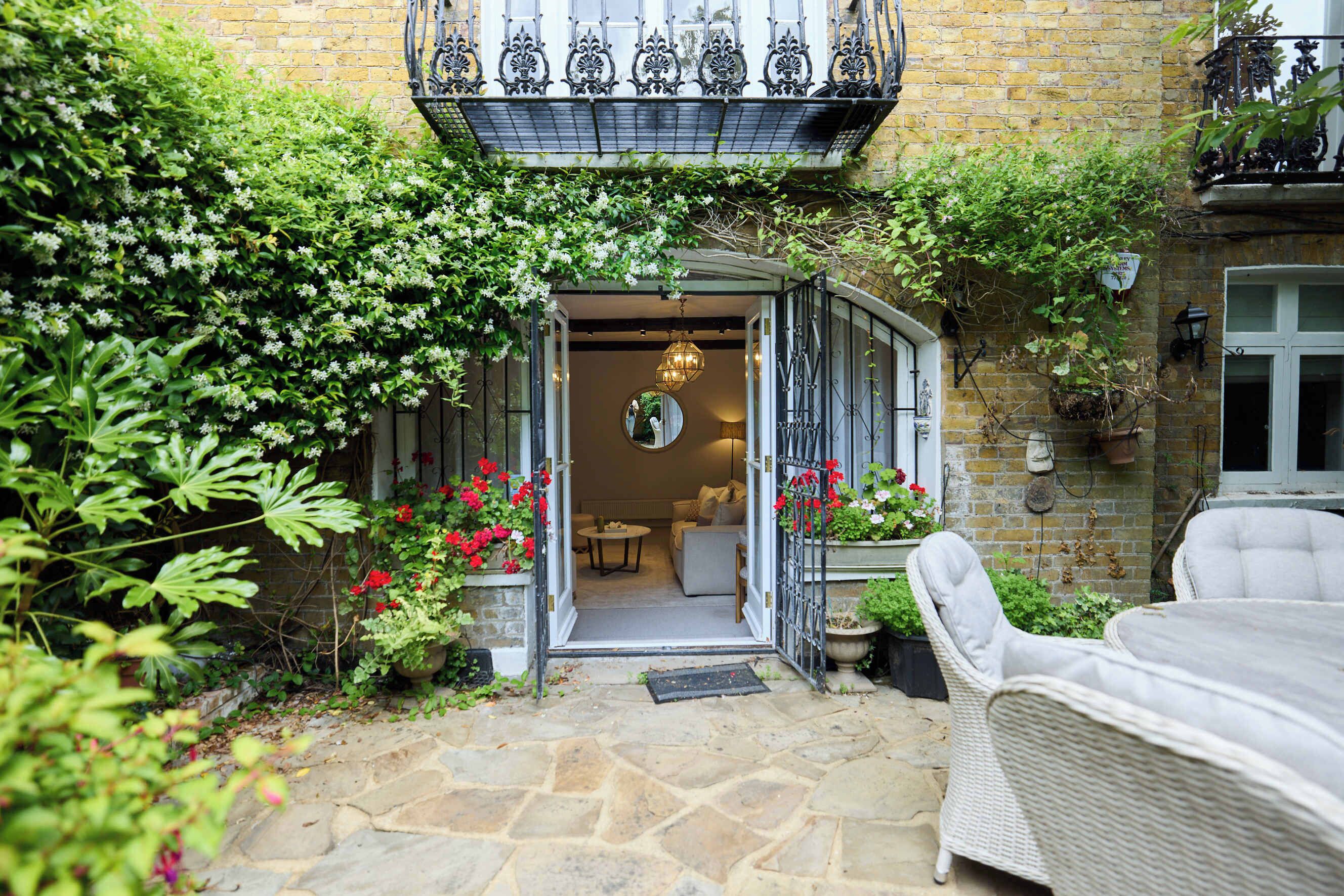 A stone-paved entrance to a building with a balcony above, surrounded by greenery and potted plants.