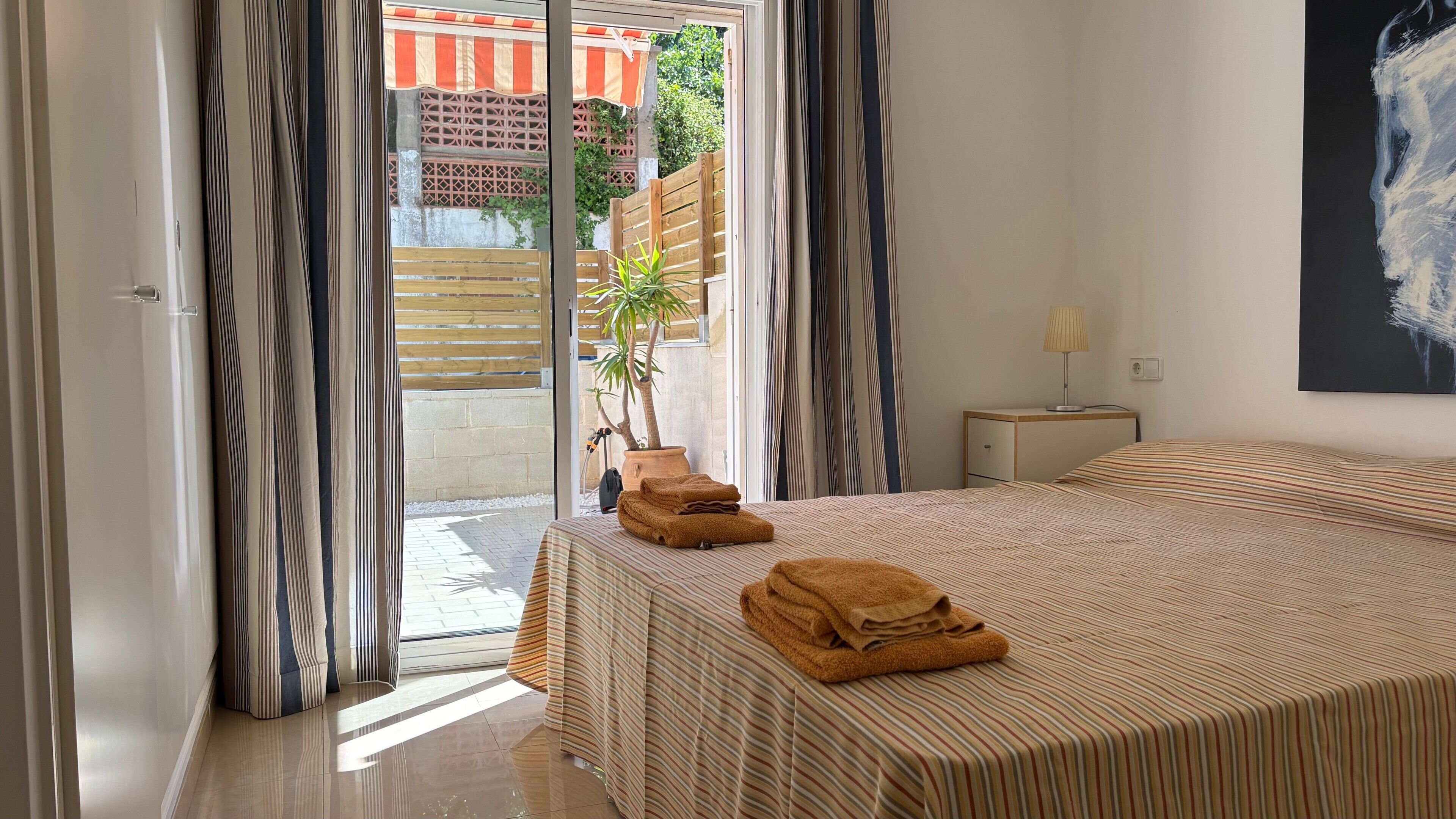 A hotel room with a bed, a nightstand, a lamp, a potted plant, and a view of the outside.