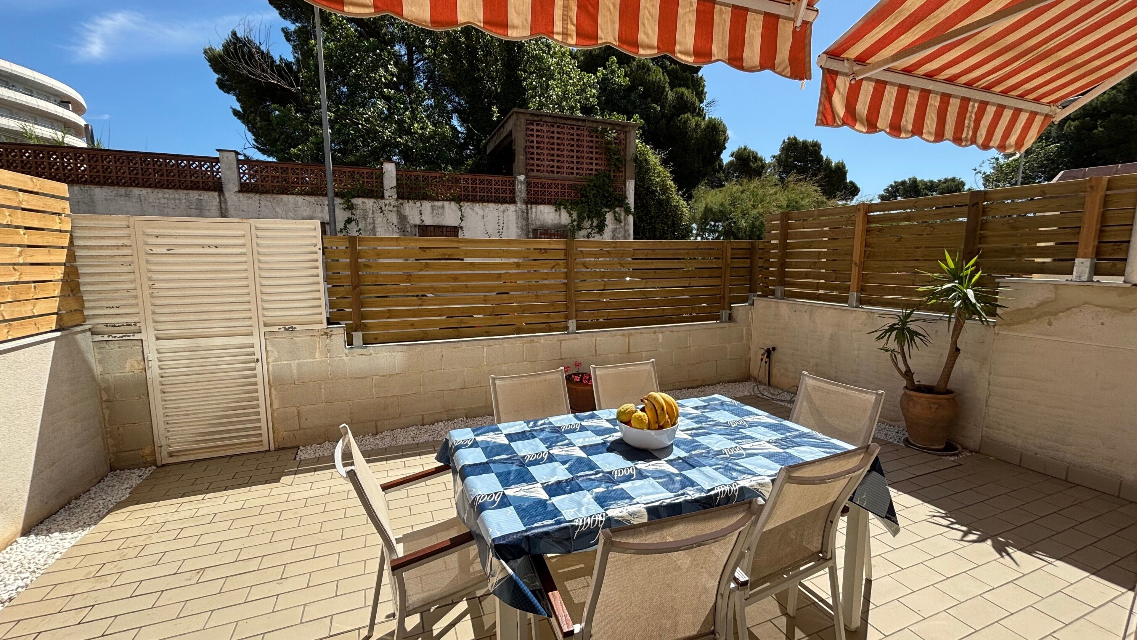An outdoor patio with a table covered in a blue and white checkered tablecloth, surrounded by chairs, and a potted plant.