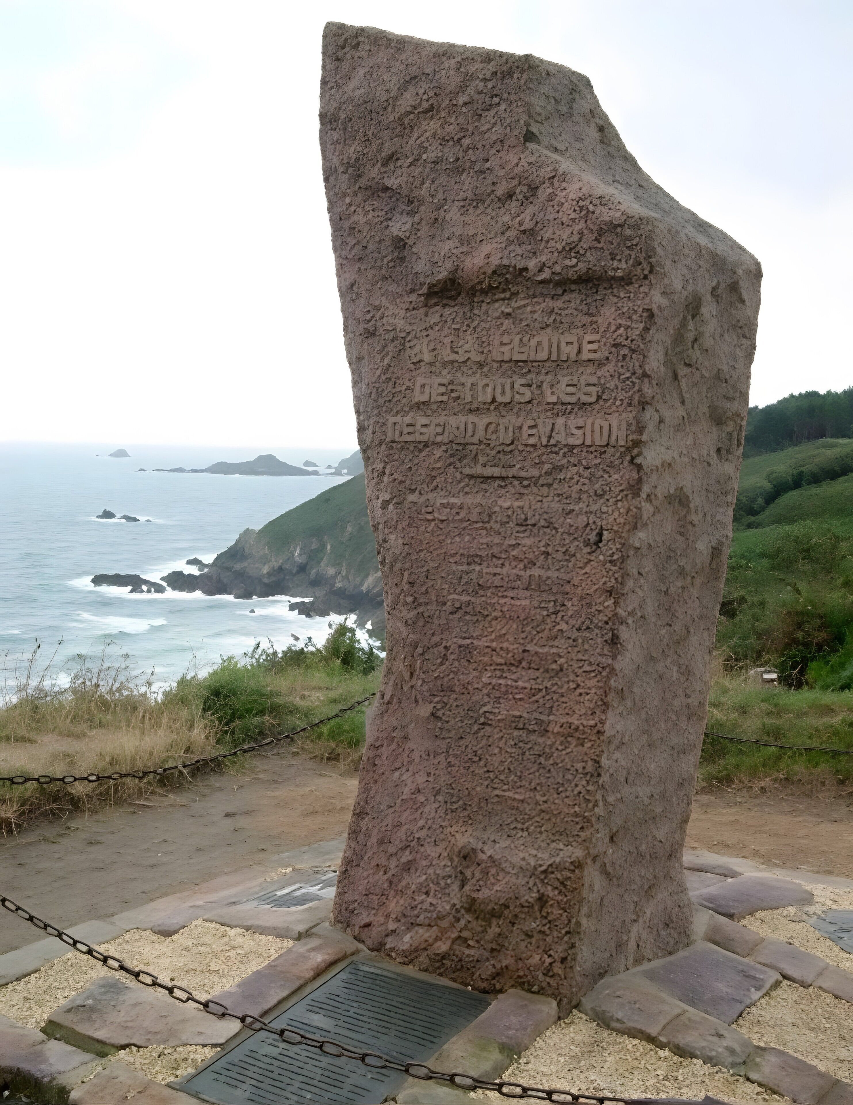 A stone monument with inscriptions, surrounded by a chain-link fence, overlooking a coastal landscape with hills and the sea.