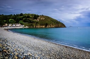 A pebble beach with a curved shoreline, a hill with buildings, and calm sea.
