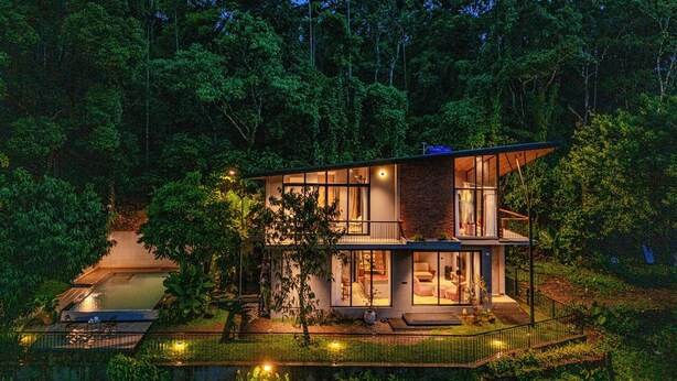 Une maison moderne à deux étages avec une piscine, entourée d’une forêt dense.