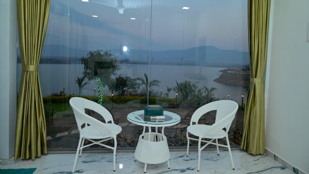A room with a large window, two white chairs, a small round table, and a view of a body of water and greenery. - Bellajooz Lake Resort (Mawal)