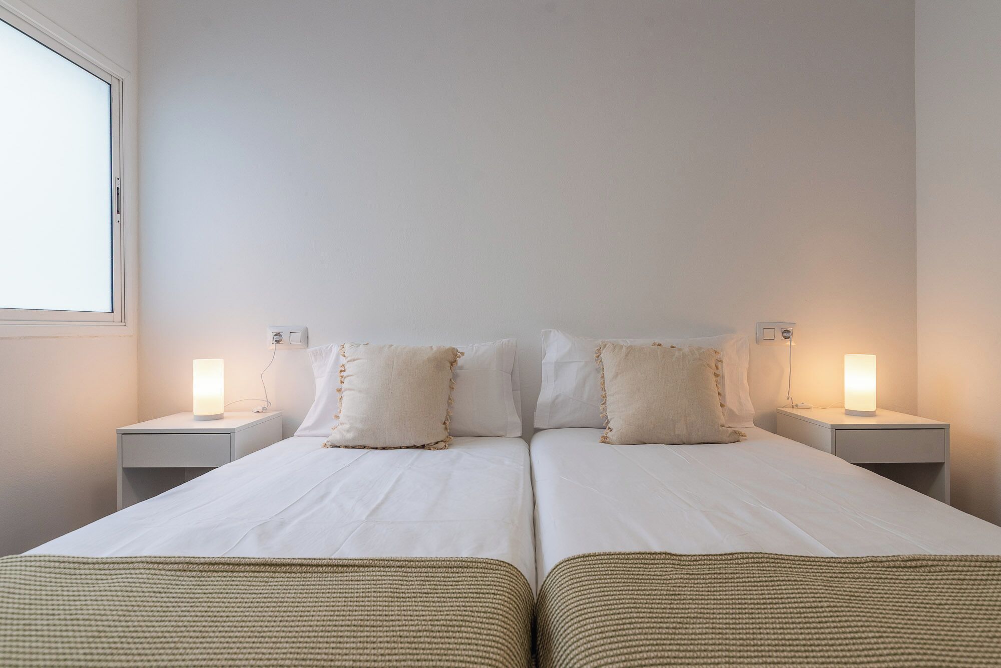 Un lit double avec deux tables de chevet, chacune équipée d’une lampe, dans une pièce aux murs unis et dotée d’une fenêtre.