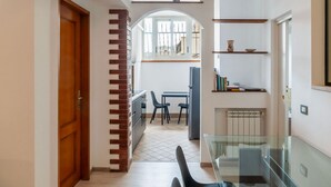 Un pasillo con una puerta de madera, pared de ladrillo, una mesa de cristal y vistas a un comedor a través de un arco.