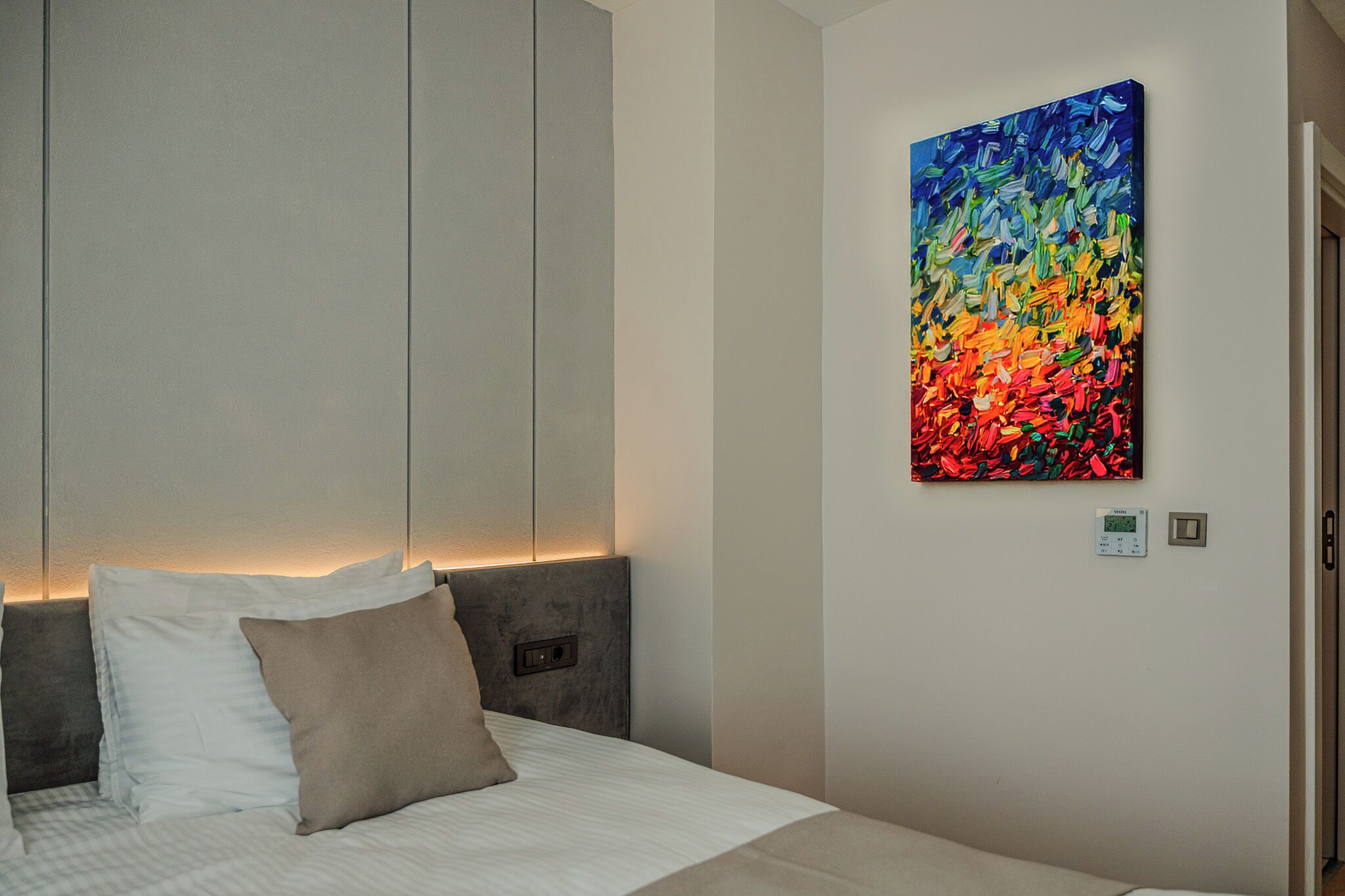 A hotel room with a bed, a headboard, a wall-mounted painting, and a thermostat.