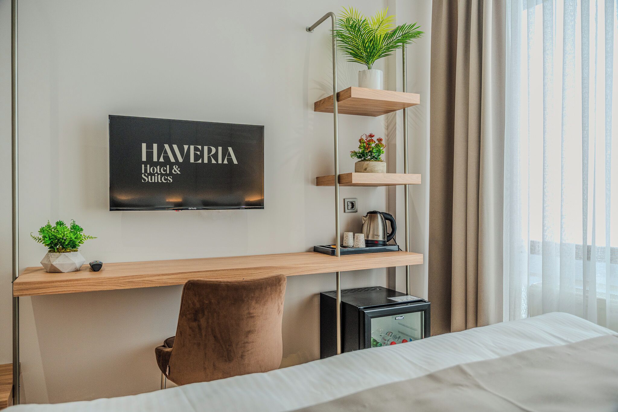 A hotel room with a bed, a desk with a computer, a coffee maker, a TV displaying 'HAVERIA Hotel & Suites', and a plant on a shelf.