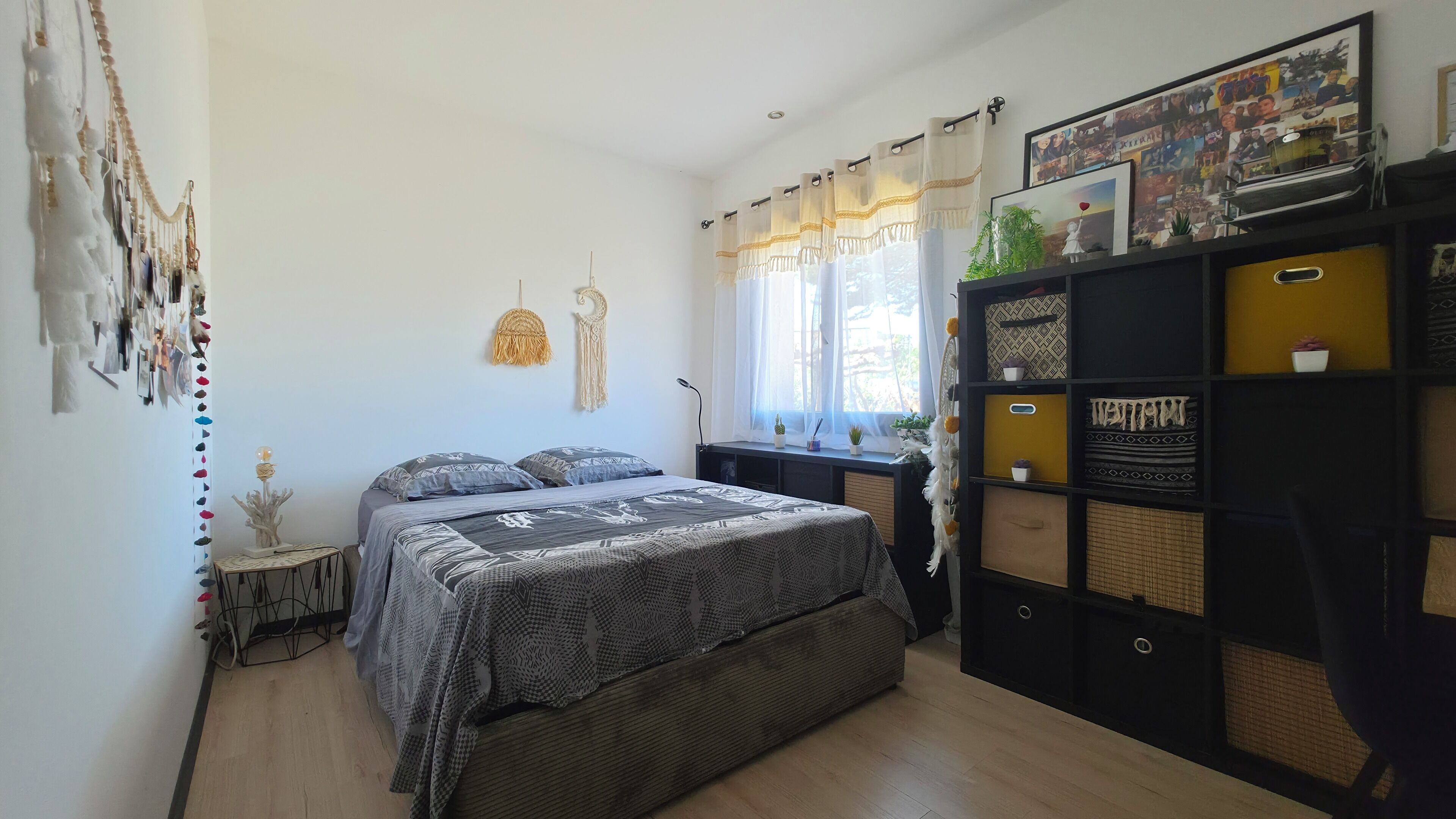 A bedroom with a bed, a bedside table, a wall-mounted shelf with decorative items, and a window with curtains.