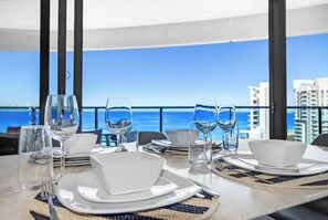 Une table à manger dressée pour quatre personnes, avec de l’assiette, des bols et des verres blancs, donnant sur une mer bleue et limpide et sur des bâtiments.