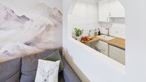 A modern living room with a mountain landscape mural, a gray sofa with a white deer pillow, and a kitchen with white cabinets and a wooden countertop visible through an archway. - Vacation apartment Hüttenzauber for 4 persons, 54 m² (Inzell)
