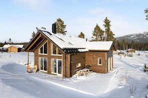 A wooden cabin with large windows, a snow-covered roof, and a clear view of the surrounding snowy landscape.