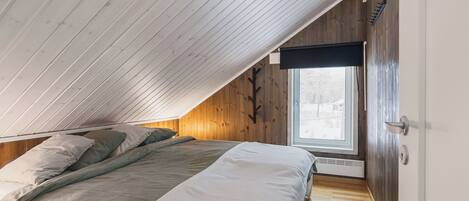 A bedroom with a sloped ceiling, wooden walls, a large window, and a bed with bedding.