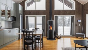 A modern kitchen with grey cabinets, a dining area with a table and chairs, a fireplace, and large windows with a snowy view. - Orrmötesvägen 21 - Funäsdalen (Funäsdalen)