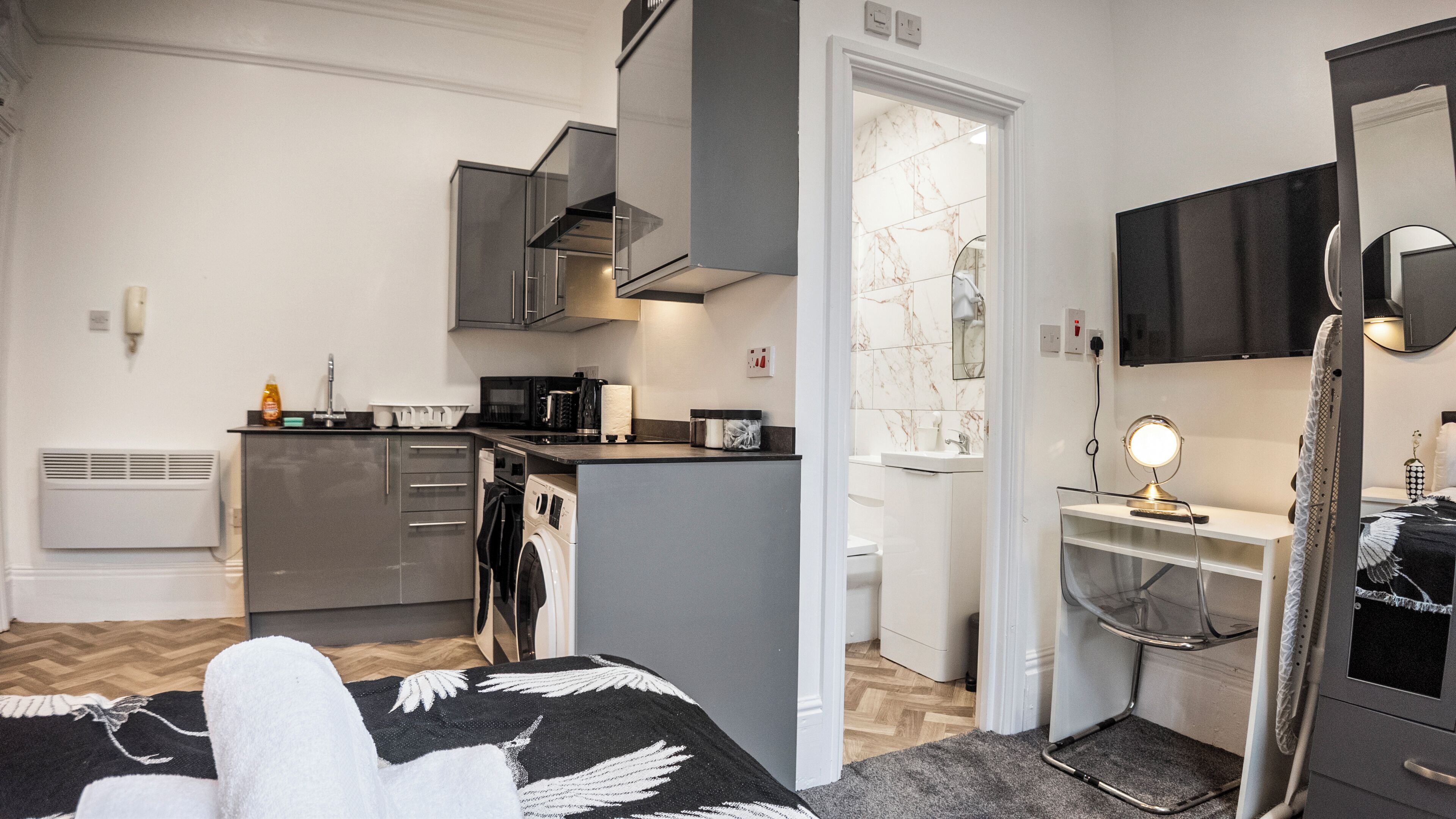Quarto de hotel pequeno com cozinha compacta, banheiro com espelho e cama com manta decorativa.
