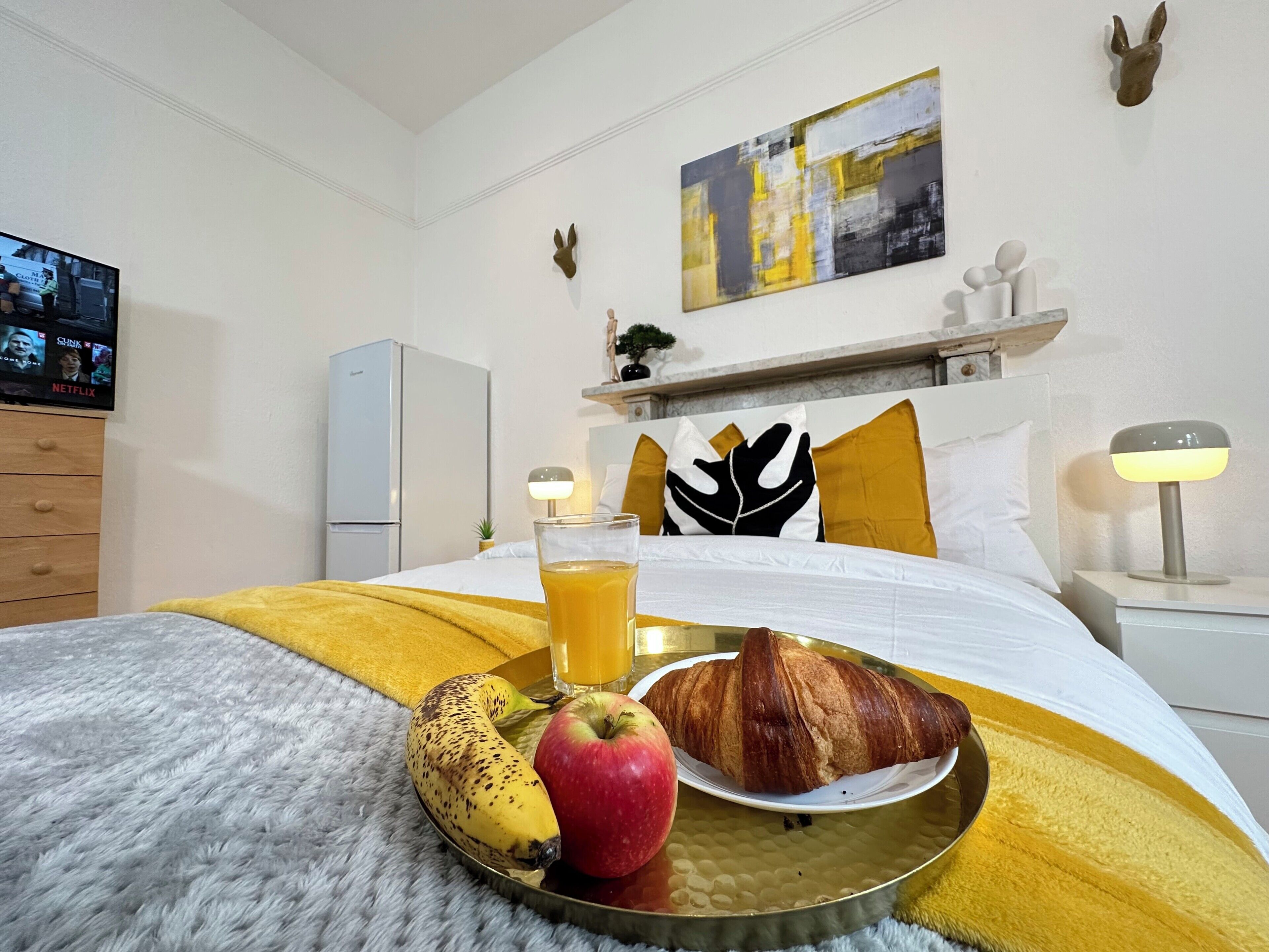 A hotel room with a bed, a bedside table with a lamp, a TV on the wall, and a breakfast tray with a banana, apple, and croissant.