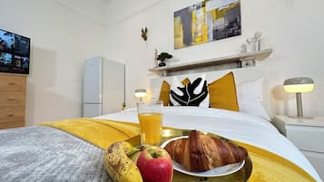 A hotel room with a bed, a bedside table with a lamp, a TV on the wall, and a breakfast tray with a banana, apple, and croissant.