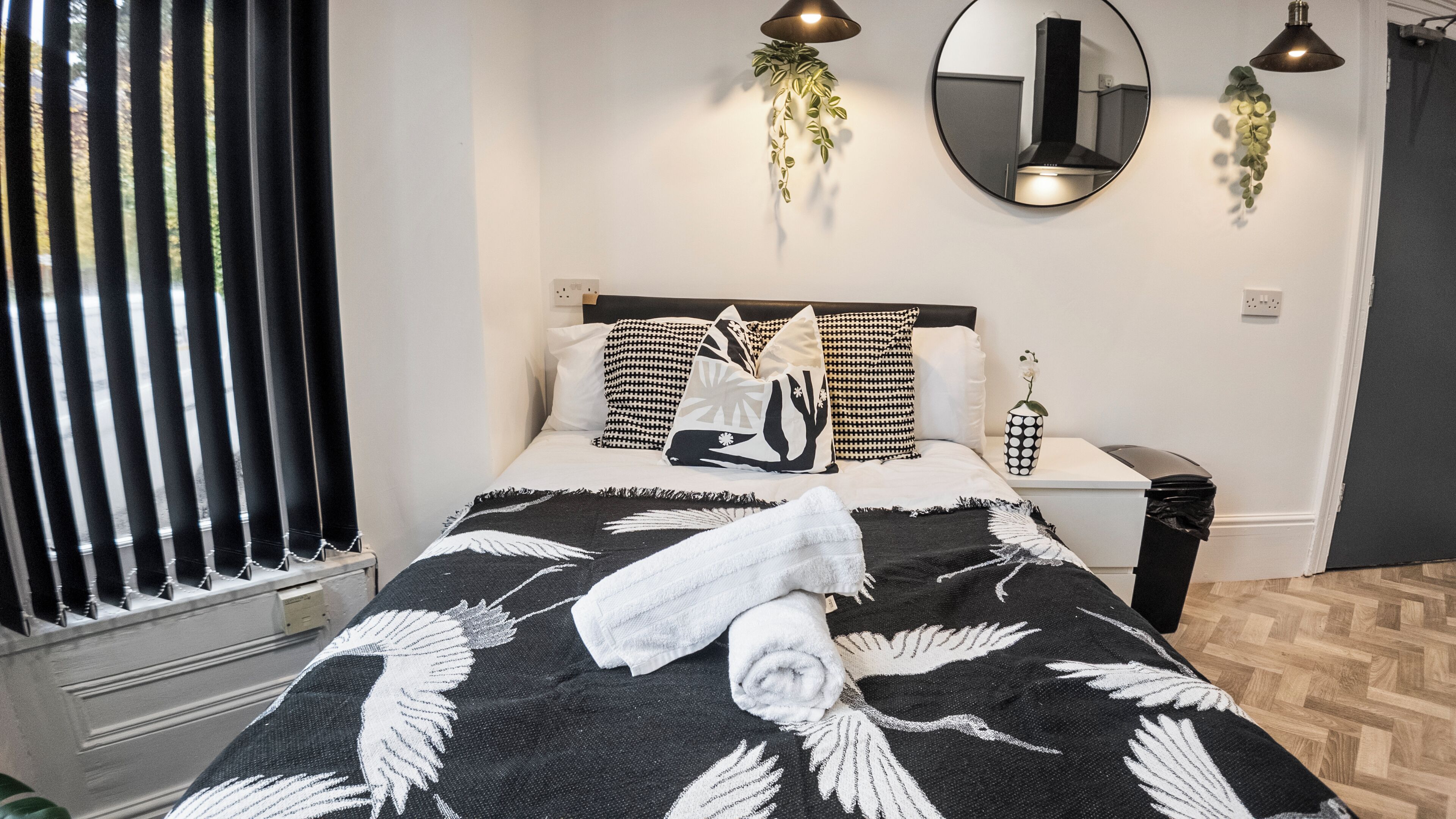 A neatly made bed with a black and white patterned bedspread, a rolled white towel, and a decorative pillow with a nature design. There are two wall-mounted lamps and a mirror above the bed.
