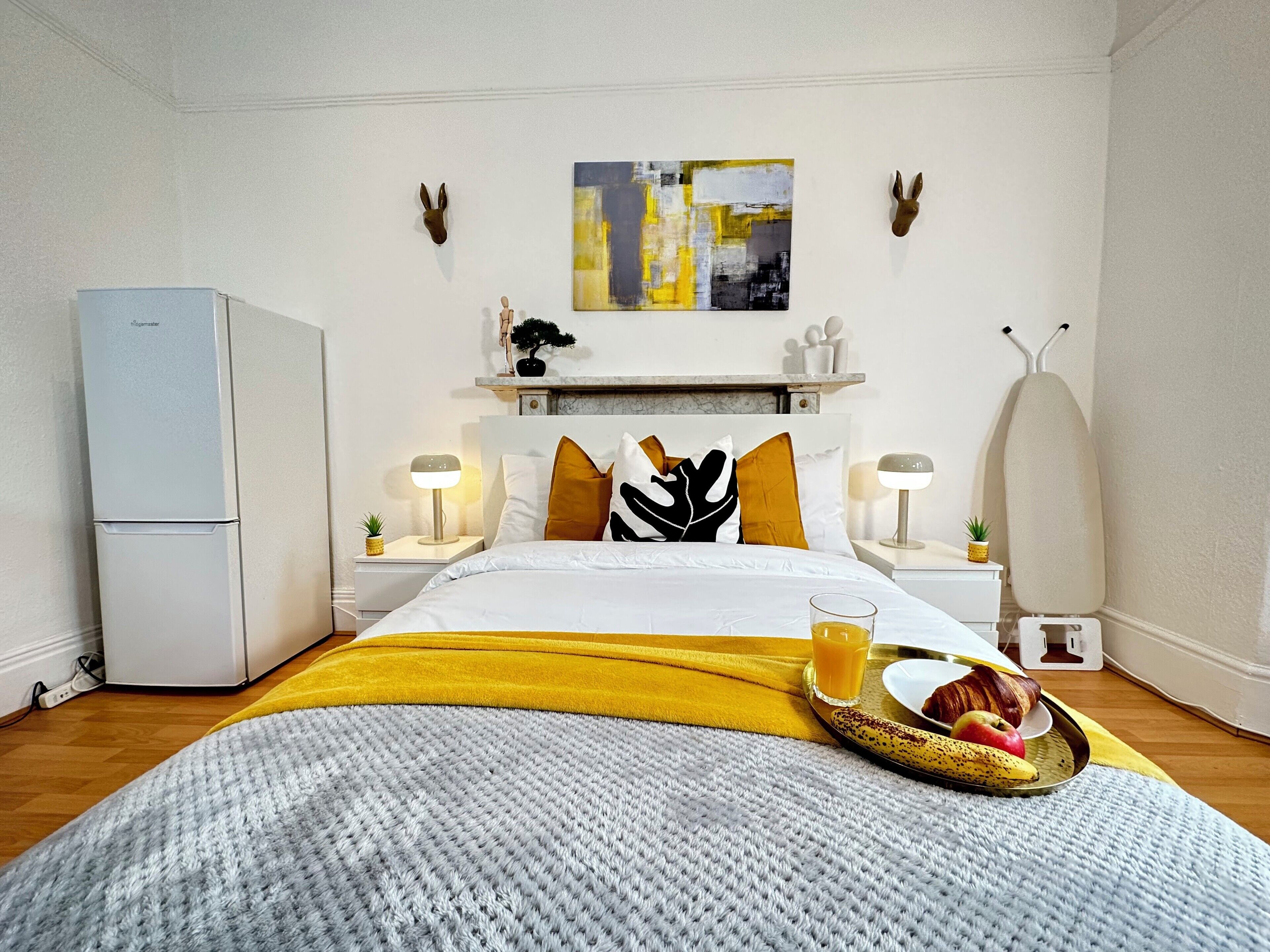 Une chambre d’hôtel comprenant un lit, une table de chevet, une lampe, un panier de fruits et une étagère fixée au mur.