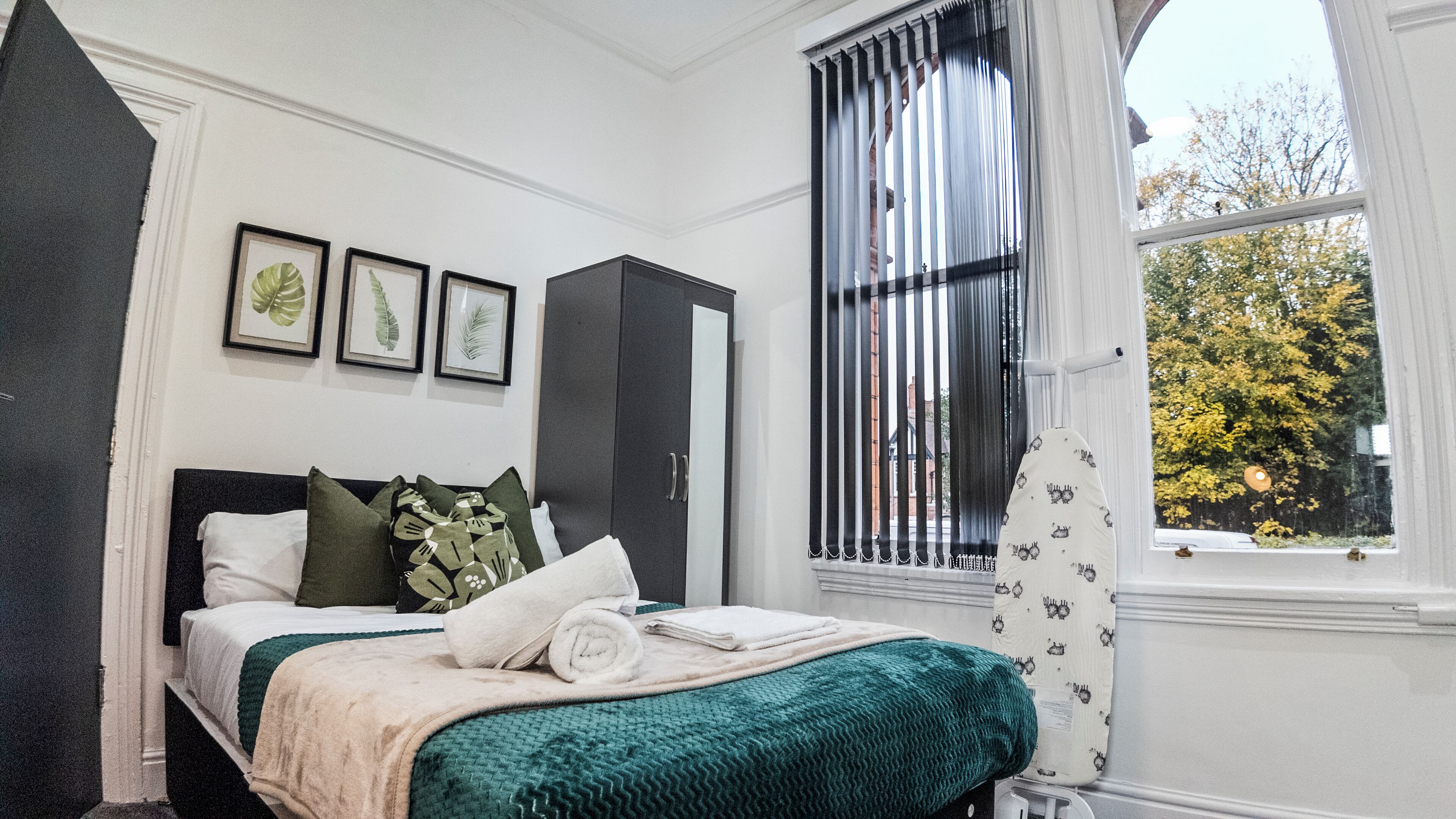 A bedroom with a bed, a wardrobe, a window with blinds, and framed leaf artwork on the wall.