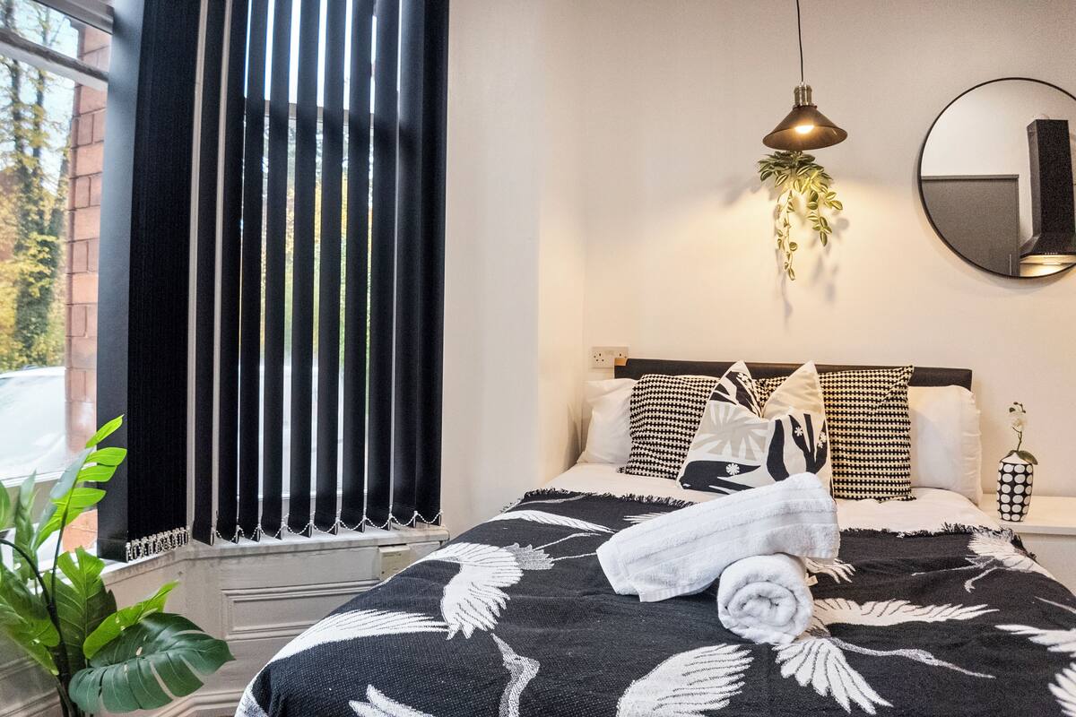 A bedroom with a bed, a window with blinds, a hanging plant, and a mirror on the wall.