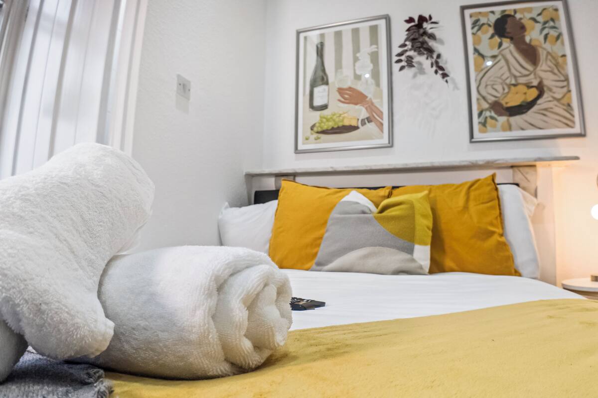 A neatly made bed with a white towel, a yellow blanket, and a remote control on the side. There are two pillows, one yellow and one gray. The room has a white wall with two framed pictures and a small table with a lamp.