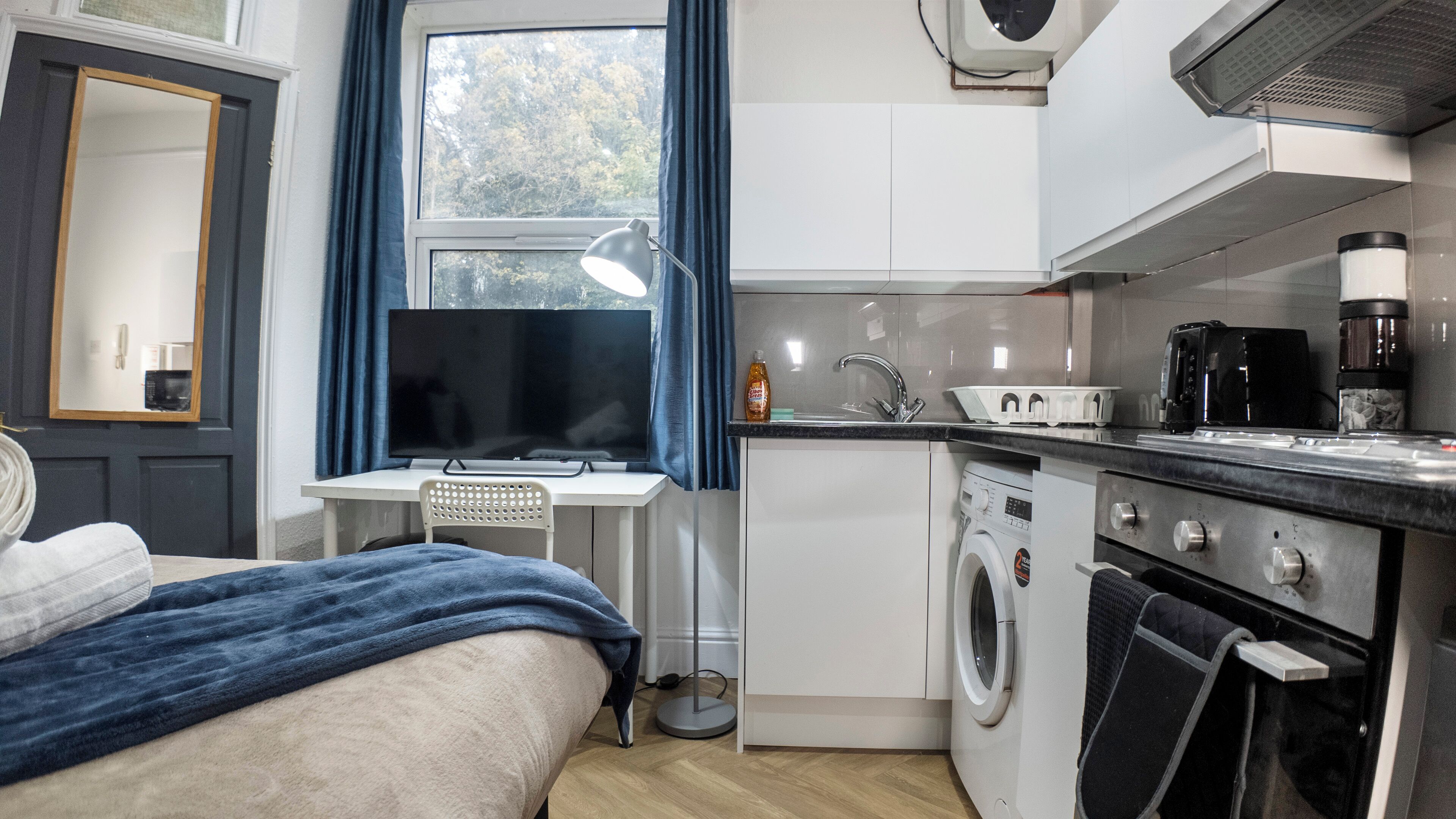 A compact hotel room with a bed, a desk with a computer, a washing machine, and a kitchenette.