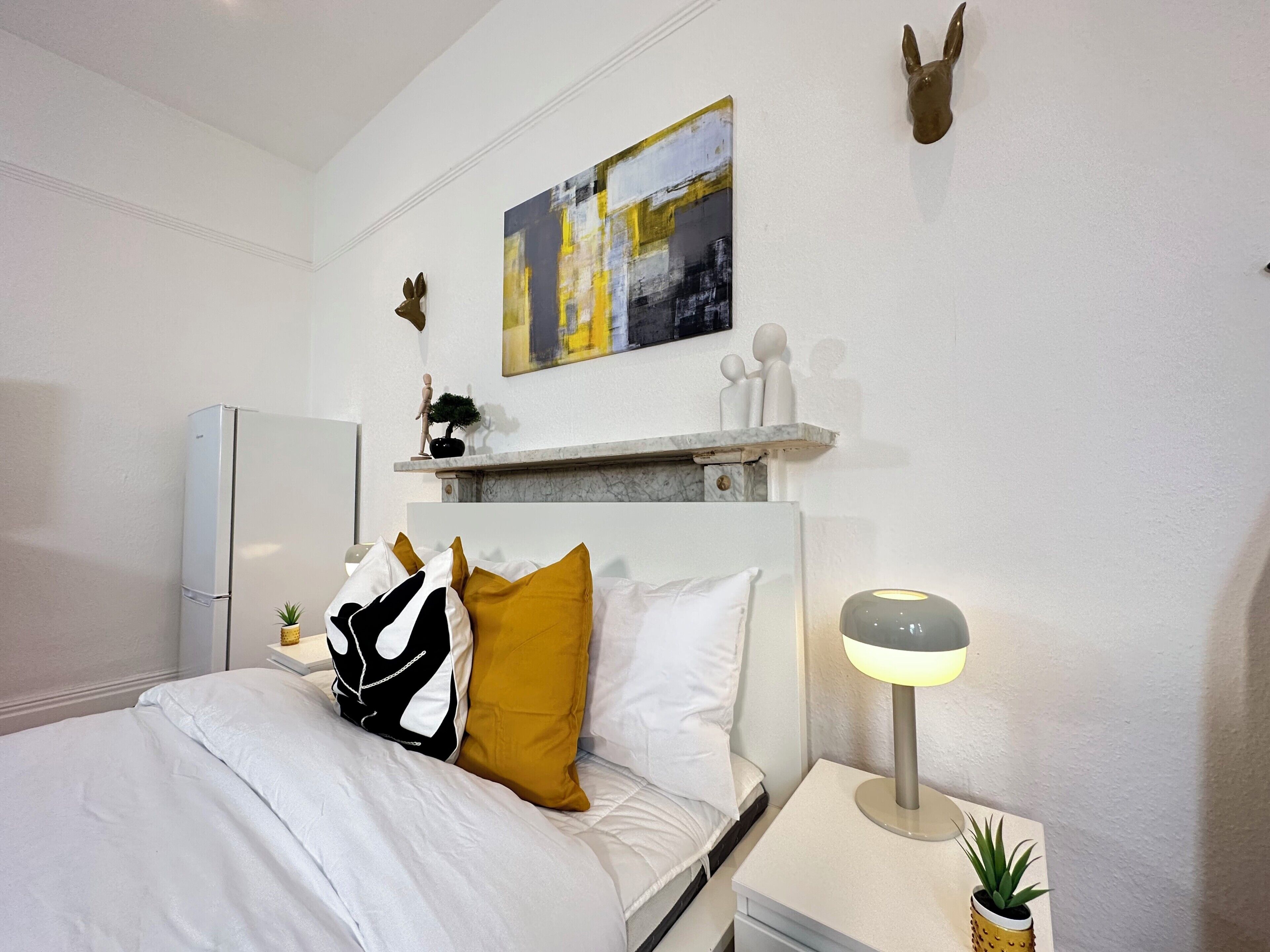 A hotel room with a bed, a nightstand with a lamp, a small plant, a wall-mounted shelf with decorative items, and a painting above the shelf.