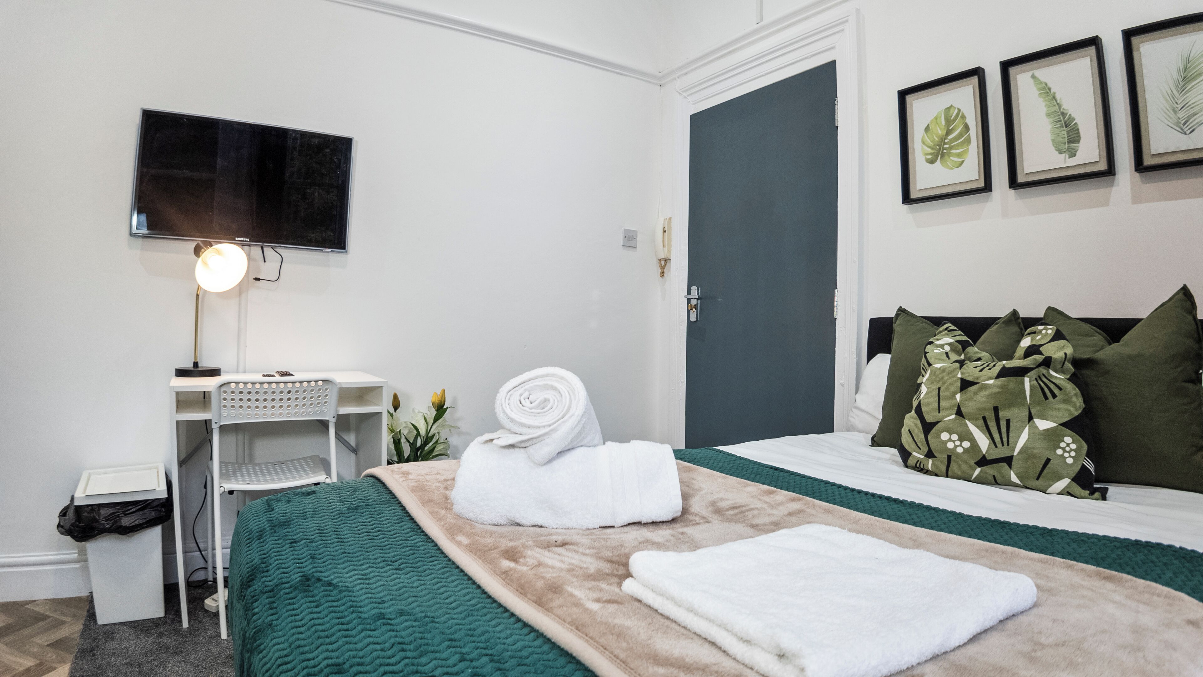 A hotel room with a bed, a desk with a lamp, a TV mounted on the wall, and framed artwork.