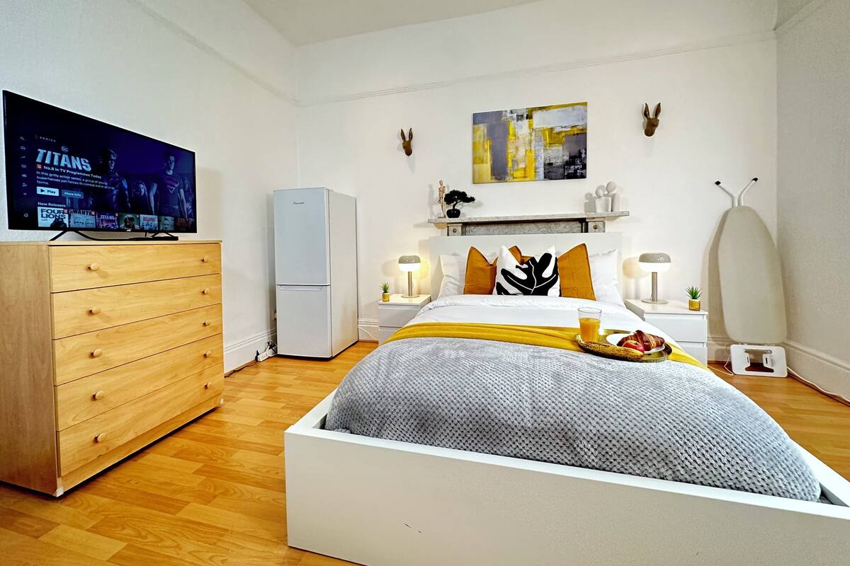 A modern hotel room with a bed, a television, a wardrobe, a small refrigerator, and a bedside table with a lamp.