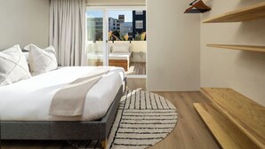 A modern bedroom with a bed, a round rug, wooden shelves, and a view of buildings through a glass door. - Magic on Main (Cape Town)