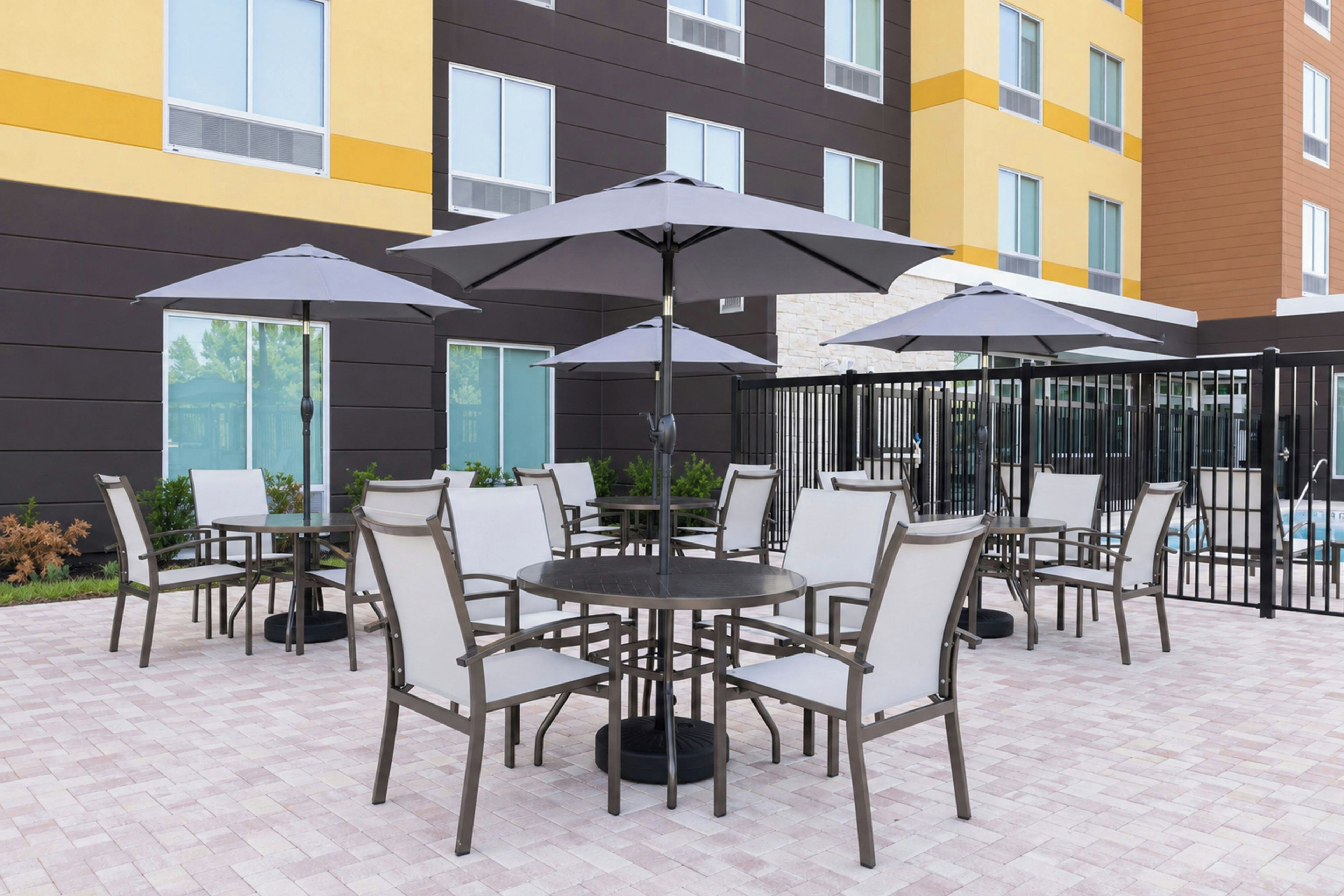 An outdoor patio area with multiple tables and chairs, umbrellas, and a pool in the background.
