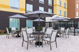 An outdoor patio area with multiple tables and chairs, umbrellas, and a pool in the background.