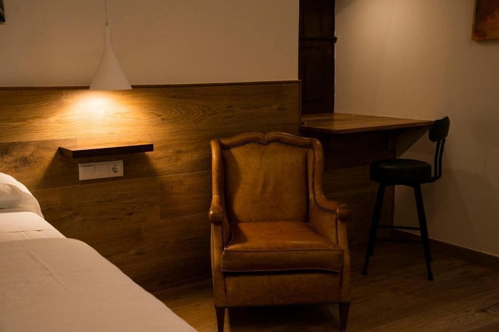 Una habitación con cabecera de madera, una silla de madera, una mesita pequeña y una lámpara colgante blanca.