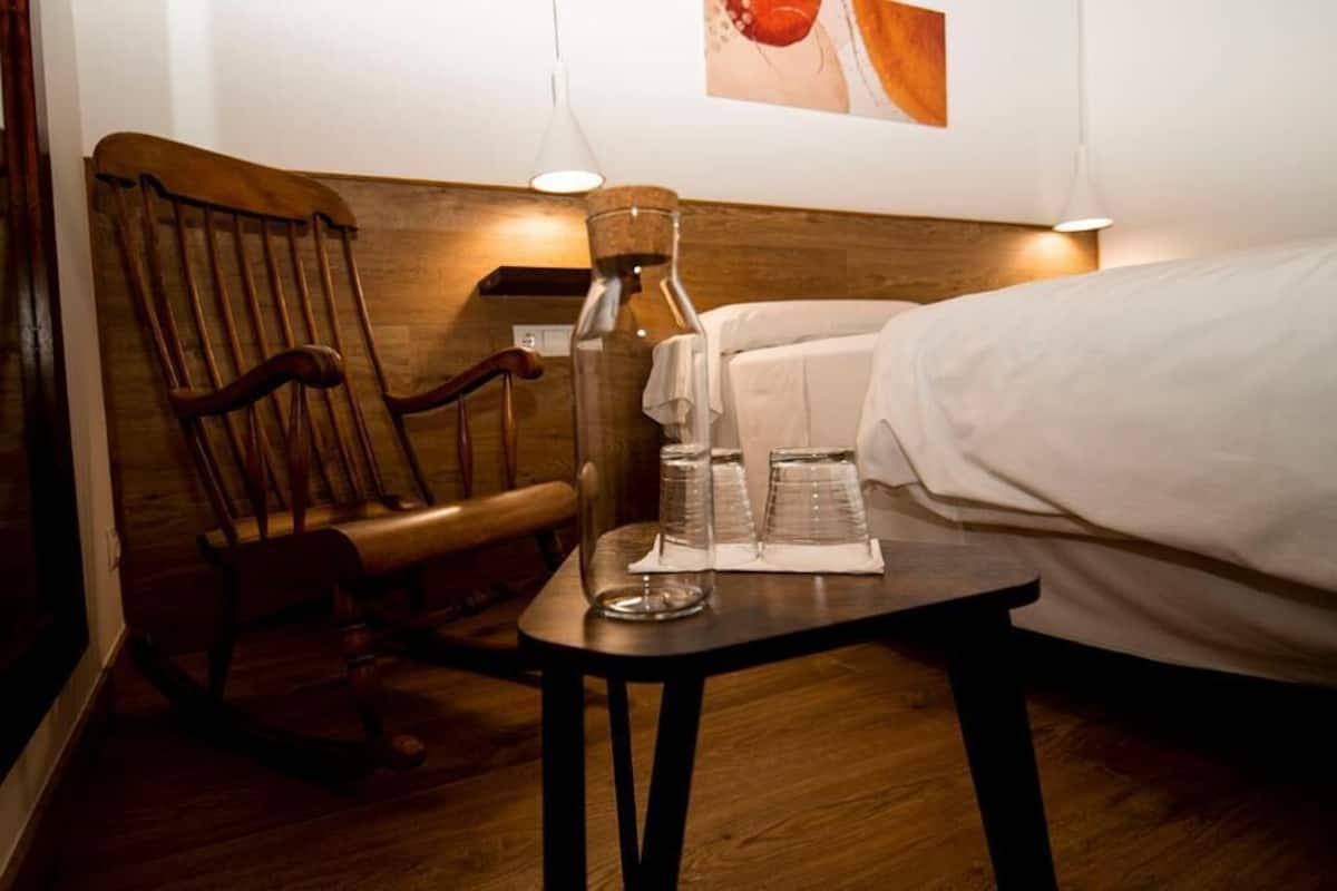 Una silla de madera, una mesa pequeña con vasos y una botella, y una cama en una habitación con suelo de madera y pared blanca.