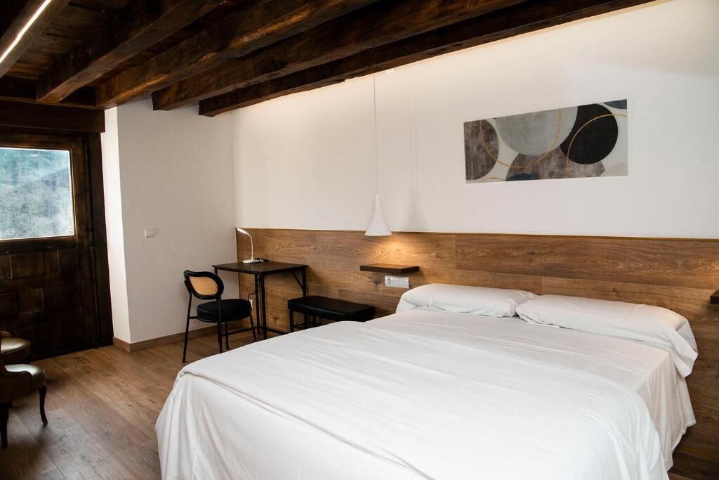 Un dormitorio con techo de madera, cama con ropa de cama blanca, escritorio con silla y una lámpara de pared.